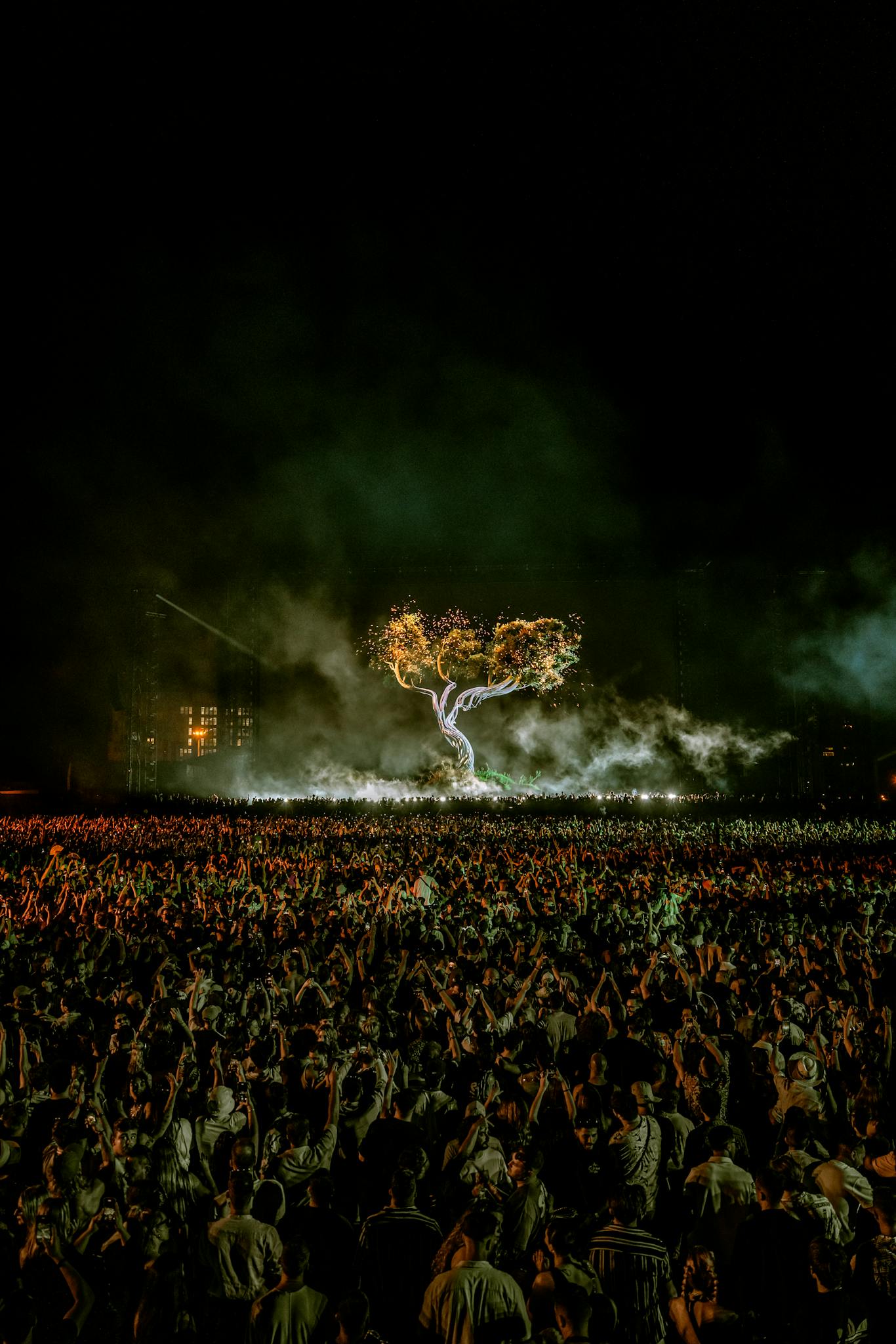 A massive crowd enjoys a night concert with a spectacular illuminated tree on stage, lit by vibrant lights.
